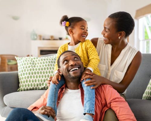 Cheerful little girl sitting on father shoulder while playing with mother at home. Happy black  family enjoying weekend at home. Cute little daughter sitting on fathers shoulder and play with her mom.
